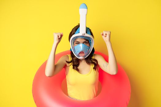 Portrait of enthusiastic young woman on vacation, wearing snorkling, diving mask and swimming ring, standing over yellow background.