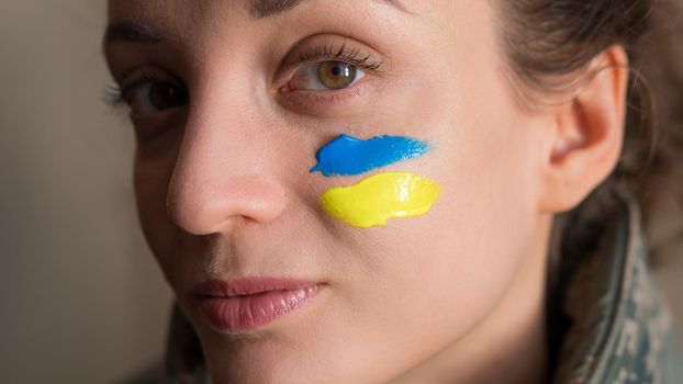 Indoor portrait of young girl with blue and yellow ukrainian flag on her cheek wearing military uniform, mandatory conscription in Ukraine, equality concepts.