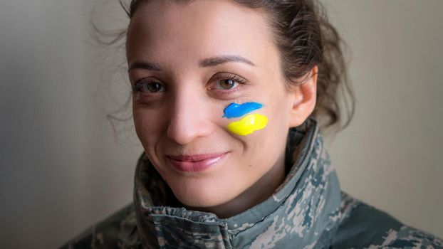 Indoor portrait of young girl with blue and yellow ukrainian flag on her cheek wearing military uniform, mandatory conscription in Ukraine, equality concepts.