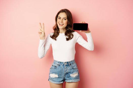 Stylish young woman introducing company website, mobile app, showing smartphone horizontally and peace sign, smiling happy, standing over pink background.