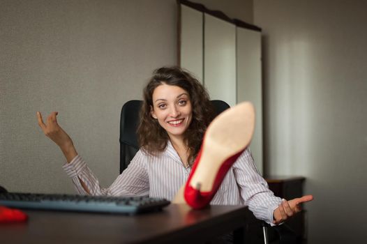Portriat of brunette with legs on office table with laptop, hot woman in red shoes on high heels sitting at the desk. Concept of seductive secretary, female fashion, relax at work.