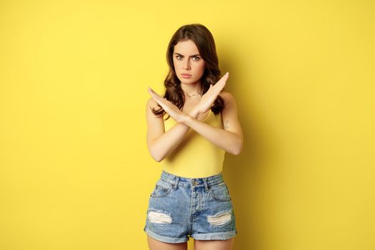 Serious stylish woman showing stop, rejection, prohibition gesture, decline something, disapprove, standing over yellow background.