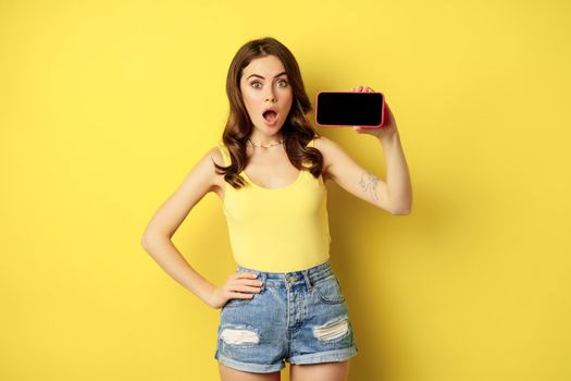 Shocked and amazed stylish girl, showing smartphone screen, app on horizontal mobile phone display, standing in summer clothes over yellow background.