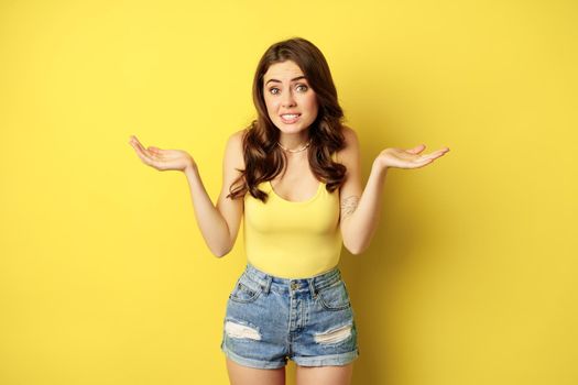 Woman shrugging shoulders and looking clueless, apologizing and smiling awkward, dont know, puzzled, has no idea, standing against yellow background.