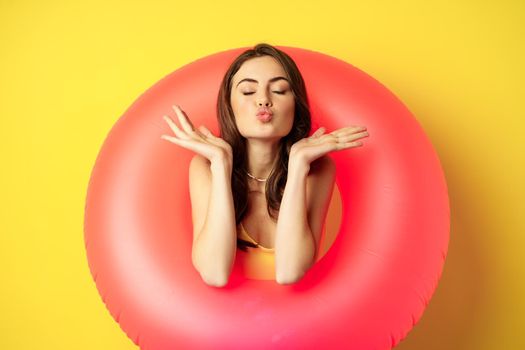 Beautiful coquettish woman inside pink swimming ring, posing on summer vacation, beach holiday concept, standing against yellow background.