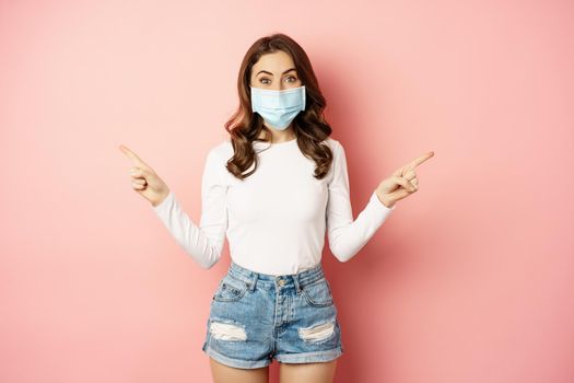 Portrait of beautiful woman in face medical mask pointing sideways, showing two choices, ways, sides, demonstrating advertisement, pink background.