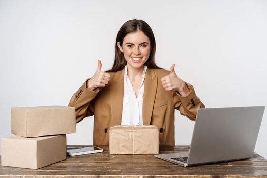 Portrait of businesswoman sitting at table, packing boxes with orders for clients, working on laptop, showing thumbs up, posing against white background.