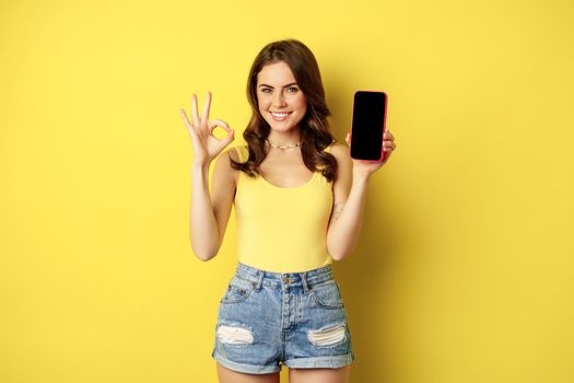 Pretty brunette woman, showing mobile phone screen, smartphone app interface and okay sign, recommend website, store, standing against yellow background.