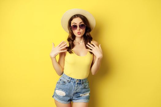 Stylish girl in straw hat and tank top, ready for summer, going on vacation and smiling pleased, standing over yellow background.