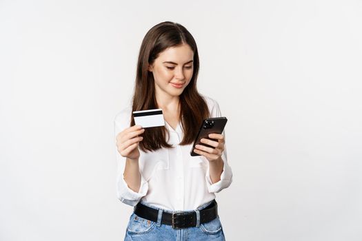 Portrait of young woman looking at smartphone screen, paying for purchase online with credit card, standing over white background, placing order.