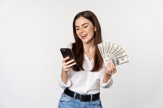 Portrait of happy smiling woman using mobile phone app, holding money cash, standing over white background. Copy space