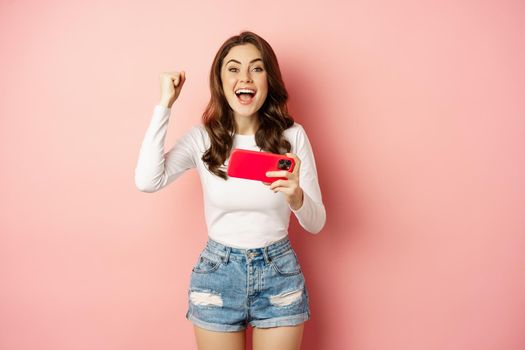 Beautiful girl looking enthusiastic, scream of joy and excitement, winning on mobile phone video game, watching smth on smartphone and celebrating, pink background.