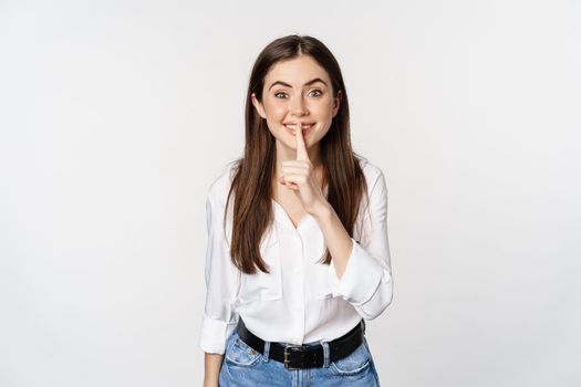 Smiling beautiful woman shushing, making hush shh sign, press finger to lips and winking, hiding secret, standing in blouse and jeans over white background.