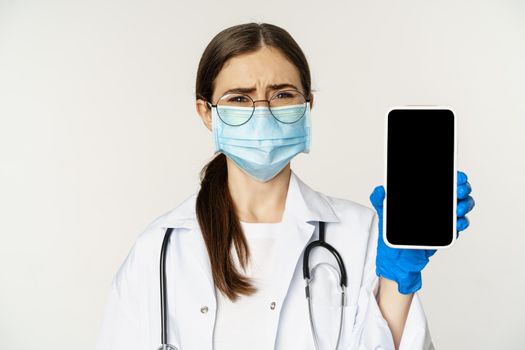 Online medical help concept. Sad and gloomy young doctor in face mask, showing smartphone screen with upset an disappointed expression, white background.
