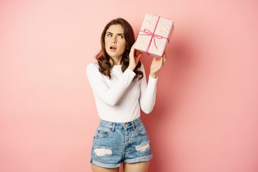 Cute smiling girl guessing whats inside pink present box, shaking gift and laughing, standing against studio background. Copy space