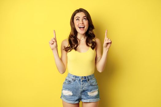 Portrait of cheerful girl smiling, pointing fingers up, showing promo text, summer advertisement, standing against yellow background.