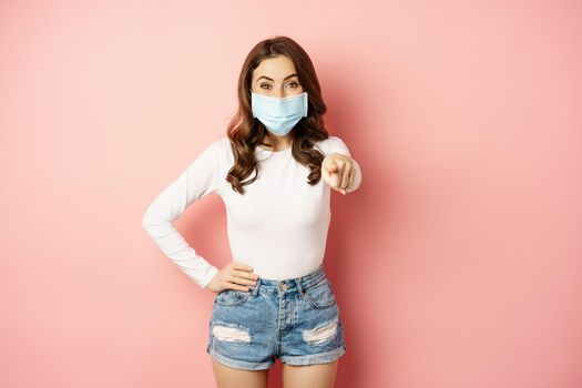 Its you. Happy smiling woman in medical face mask, pointing finger at camera, congratulating, praising, inviting people, seeing smth, standing over pink background.