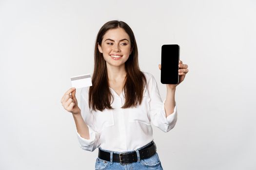 Portrait of female model showing credit card with smartphone screen, recommending application, standing over white background.