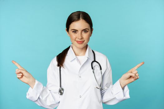 Enthusiastic woman doctor, medical worker in white coat, pointing fingers sidweays, left and right, showing health clinic promo, standing over torquoise background.
