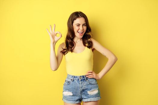 Very good, okay. Smiling coquettish woman showing ok, satisfactory sign gesture, pleased by smth, recommending, standing over yellow background.