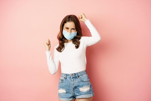 Enthusiastic brunette girl in medical face mask, dancing and laughing, celebrating victory, triumphing, winning smth, standing over pink background.