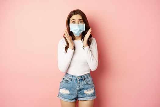 Covid-19 and quarantine concept. Stylish girl in medical face mask, looking worried and shocked at camera, standing against pink background.