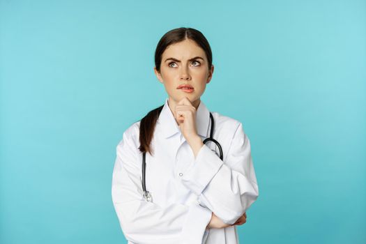 Young female doctor, hospital worker in white coat, thinking and looking away thoughtful, searching solution, standing over toquoise background.