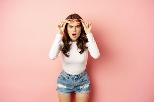 Portrait of shocked woman takes off sunglasses, looking confused, dont understand smth strange, reacting frustrated, standing over pink background.