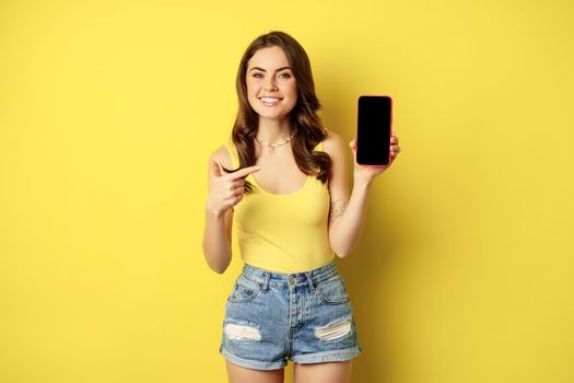 Young stylish woman showing mobile phone screen, pointing at smartphone display, app interface or online store, smiling pleased, standing over yellow background.