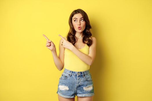 Portrait of stylish summer girl, brunette woman showing way, pointing fingers left, advertising, standing over yellow background. Copy space