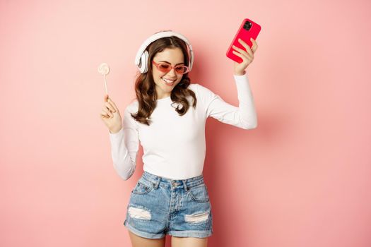 Dancing woman in sunglasses, listening music in headphones, holding lolipop and smartphone, laughing and smiling happy, standing over pink background.