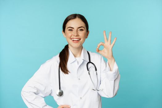 Portrait of satisfied, smiling medical worker, woman doctor showing okay, ok, zero no problem gesture, excellent sign, standing pleased over torquoise background.