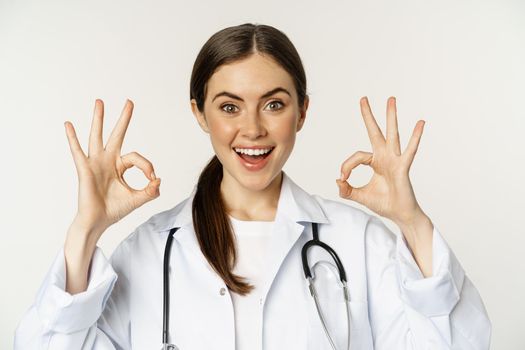 Happy beautiful woman doctor, showing okay, ok excellent sign, smiling amazed, approve smth, praise, standing in white lab coat over white background.