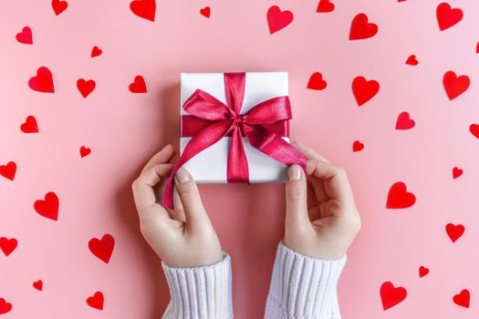 Female Hands in sweater holding a gift in white wrapping paper on pink background with red hearts. St. Valentines Day, love, tenderness, friendship and care concept. Cozy, festive, romantic wallpaper