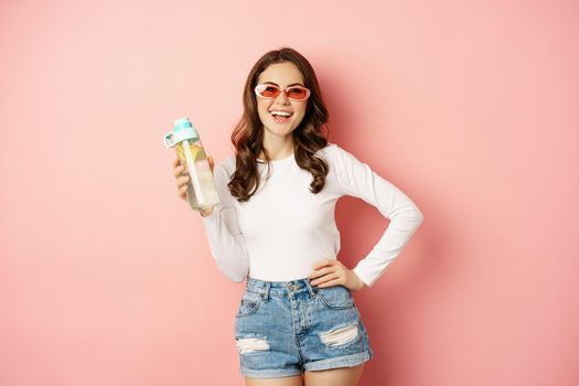 Stylish girl in spring outfit, wearing sunglasses, holding water bottle with lemon, healthy drink, laughing and smiling, standing over pink background.