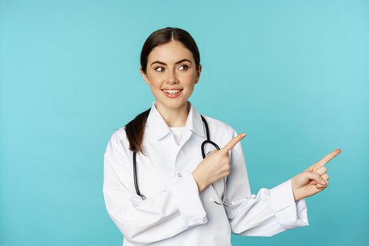 Portrait of young professional doctor, woman hospital worker, pointing fingers right, showing logo, clinic promo deal, standing over torquoise background.