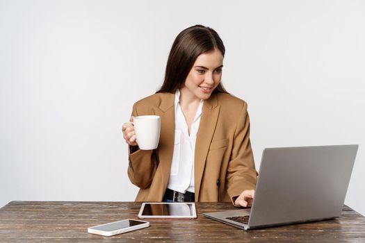 Portrait of office woman, ceo entrepreneur working on laptop, drinking coffee and answer clients on website, standing over white background.