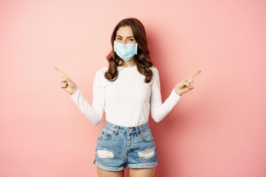 Portrait of beautiful woman in face medical mask pointing sideways, showing two choices, ways, sides, demonstrating advertisement, pink background.