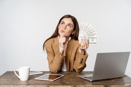 Portrait of businesswoman sitting in office with money, working and making profit income, posing happy against white studio background.