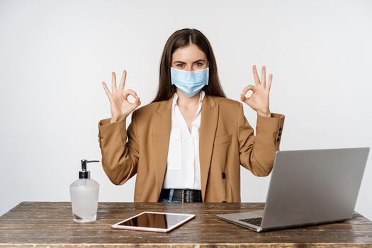 Workplace and covid-19 concept. Smiling businesswoman in face medical mask, showing okay sign after cleaning hands with sanitizer, white background.