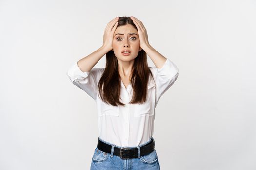 Worried and shocked young woman, looking stunned at disaster, holding hands on head, panicking, standing over white background.