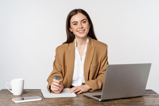 Image of businesswoman working in office, sitting at table with laptop, writing documents, posing in formal suit over white background.