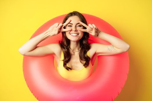 Portrait of attractive modern girl inside trendy pink beach swimming ring, showing peace v-sign and smiling happy, standing over yellow background.