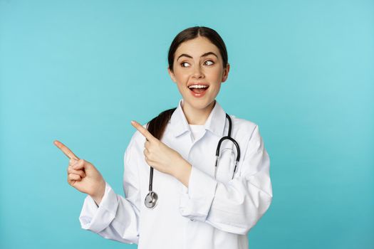 Portrait of smiling medical worker, girl doctor in white coat with stethoscope, pointing fingers left, showing medical clinic advertisement, torquoise background.