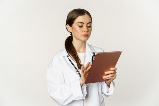 Doctor in white coat using digital tablet, reading medical data on gadget, working in hospital, standing over white background.