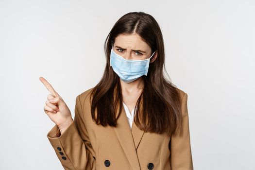 Coronavirus and people concept. Sad and concerned woman in office clothing and medical face mask, sulking upset, pointing finger at upper left corner, white background.