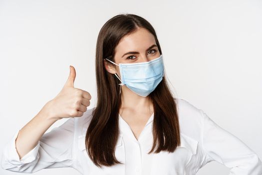 Close up portrait, face of woman in medical mask smiling, showing thumbs up, standing over white background. Copy space
