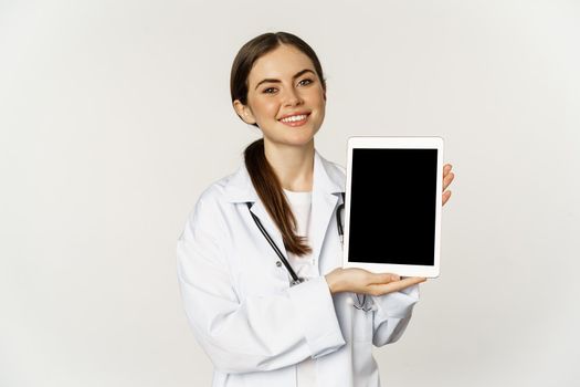 Online medical help, remote appointment. Smiling beautiful woman doctor showing digital tablet screen, demonstrating website logo, standing over white background.