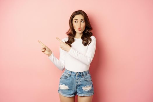 Wow, look there. Amazed feminine woman pointing fingers left, looking surprised and impressed, standing over pink background.