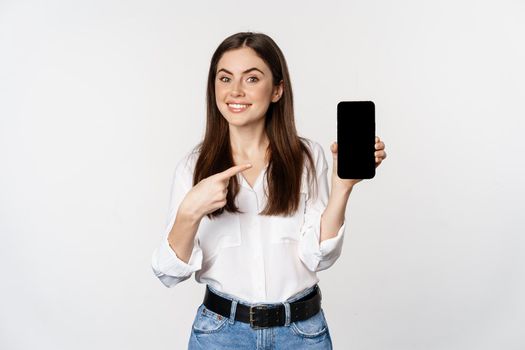 Happy beautiful woman smiling, pointing at mobile phone screen, showing advertisement, website, standing over white background.